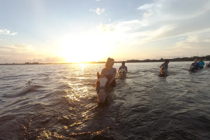people riding horses in the water at sunset